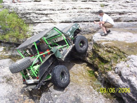 1991 Ford Bronco - A modified off-road vehicle navigates a challenging rocky stream bed, observed by a spectator.