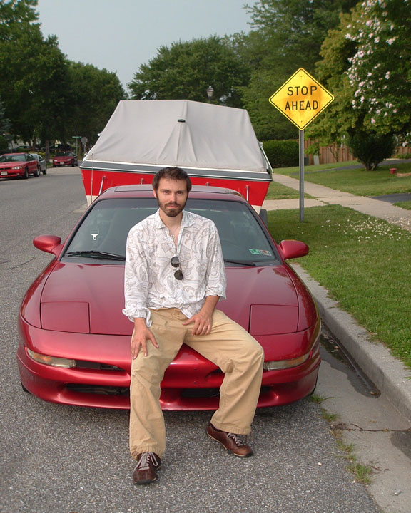 Man sitting on hood of dark red Ford Probe parked on street in front of covered boat and STOP AHEAD sign