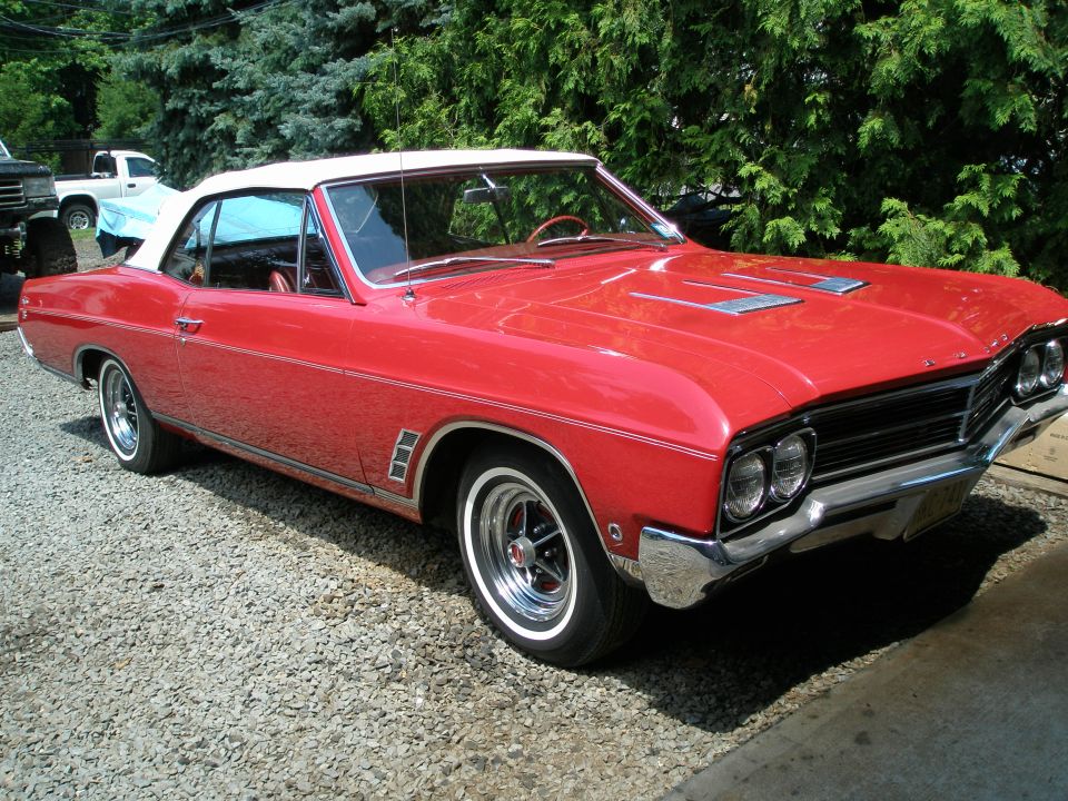 Red 1960s Buick convertible with white top parked on gravel, front 3/4 view