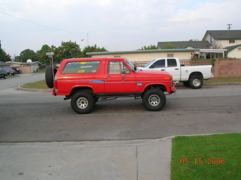1985 Ford Bronco - Side view of a red 1985 Ford Bronco with a lifted suspension and off-road tires, parked on a street next to a white pickup truck.