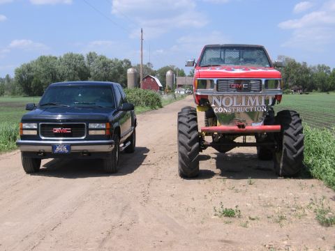 1995 GMC Sierra 1500 - Two GMC trucks, one standard and one monster truck, sit on a dirt road with a farm in the background.