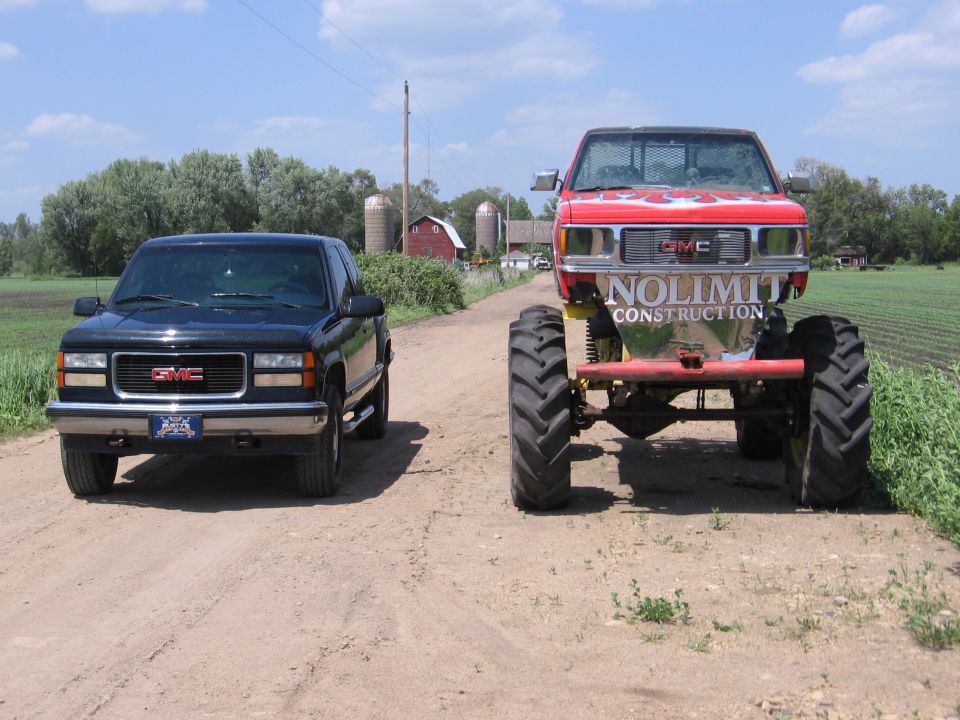 Black GMC pickup truck and a red monster truck with 'NOLIMIT CONSTRUCTION' on the tailgate, parked on a dirt road