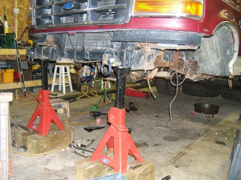 1993 Ford Bronco - The front end of a maroon Ford truck is elevated on jack stands in a garage, undergoing maintenance or repair.
