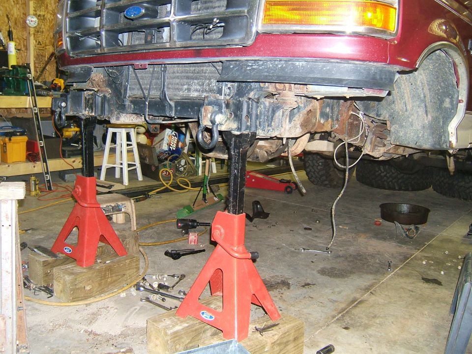 Front of a maroon Ford truck, possibly a Bronco or F-series, on red jack stands in a garage. The front bumper and some suspension components are removed