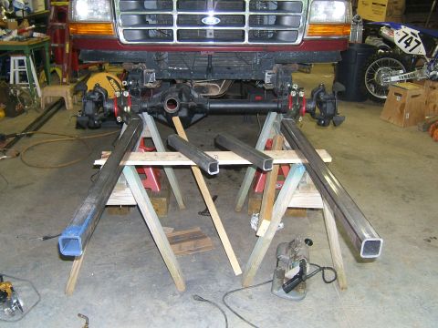 1993 Ford Bronco - The front axle of a Ford truck is suspended on sawhorses, with metal tubing and a chop saw nearby in a garage.