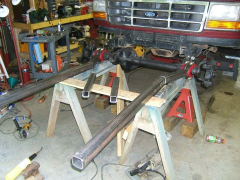 1993 Ford Bronco - Front axle assembly of a red Ford truck is supported by sawhorses in a workshop with metal tubing laid out.