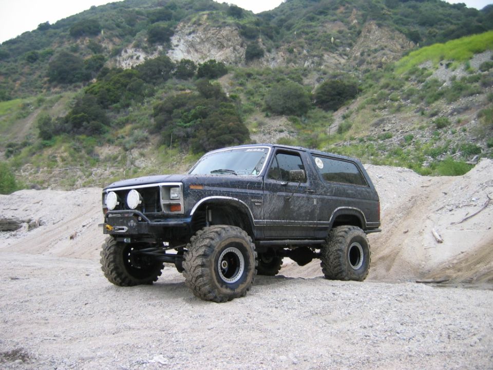 Lifted dark Ford Bronco SUV on big mud tires parked on gravel below brushy hills