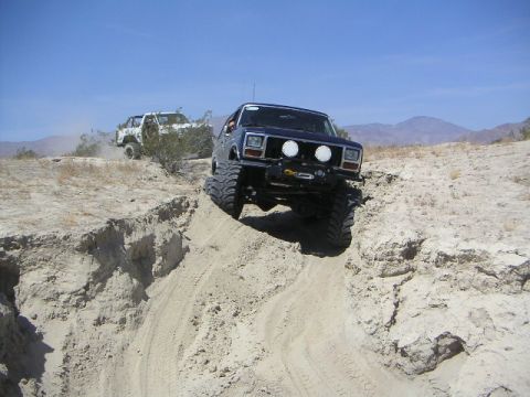 1985 Ford Bronco - Two modified SUVs navigate a challenging off-road course on a bright, clear day.