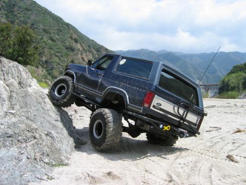 1985 Ford Bronco - A modified Ford Bronco navigates a challenging rocky terrain with a bridge visible in the background.