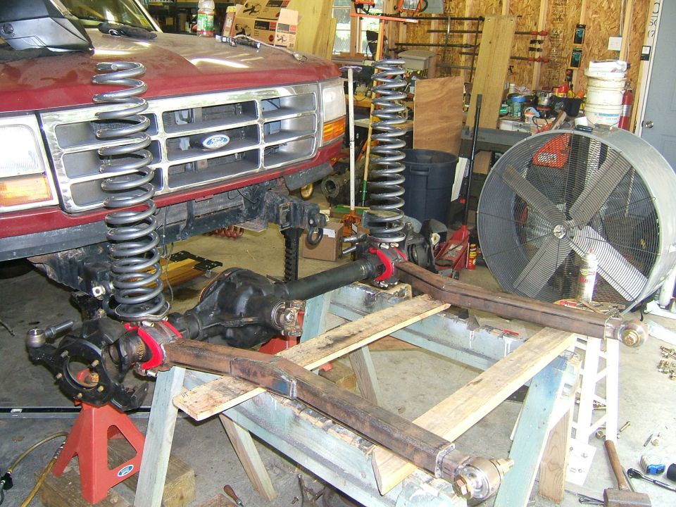 Red Ford Bronco front end on jack stands with suspension disassembled, showing large springs and axles