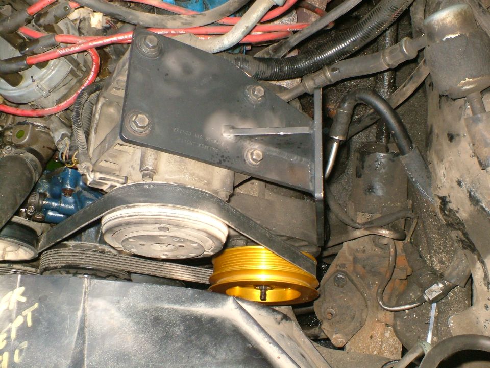 Engine bay showing an AC compressor, belts, and gold pulley. Wires and hoses are visible