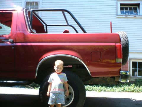 1989 Ford Bronco - A red SUV with a roll cage is parked outside a building, with a child standing beside it.