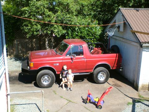 1989 Ford Bronco - A red Ford Bronco from the late 1980s is parked outside a garage, with two young children standing nearby.