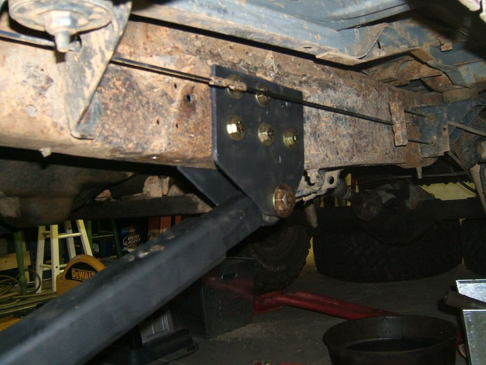 Close-up of a custom black metal suspension bracket bolted to a heavily rusted vehicle frame. A black bar extends diagonally downwards