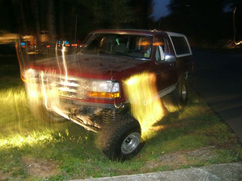 1993 Ford Bronco - A red Ford Bronco with a white top is parked on the side of the road at night, illuminated by its headlights.
