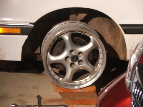 1990 Dodge Shadow - A close-up of a silver aftermarket wheel resting on wooden blocks, with a portion of a white vehicle in the background.