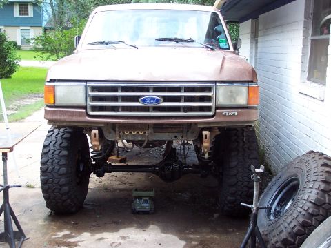 1987 Ford Bronco - A brown Ford truck with a lifted suspension and oversized tires is parked in front of a white brick building.