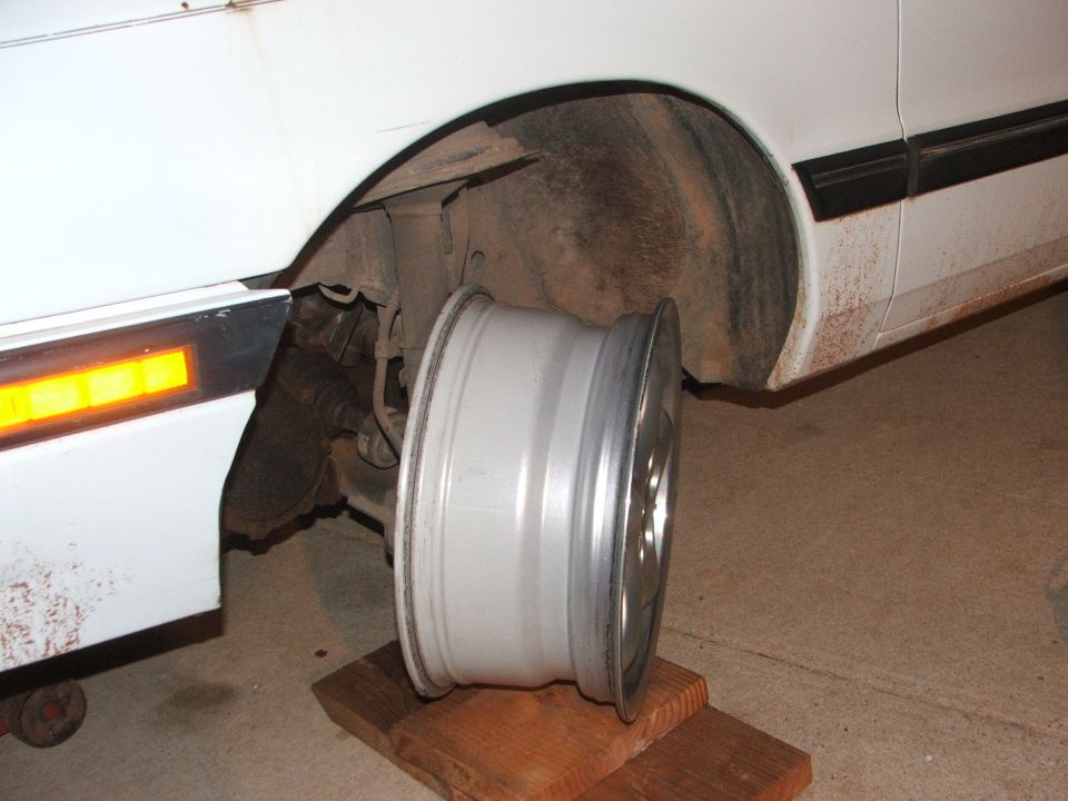 Silver aftermarket wheel resting on wooden blocks near a white car's wheel well