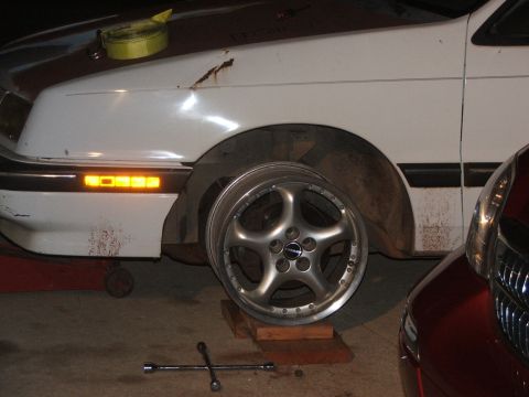 1990 Dodge Shadow - A car is on blocks with a custom wheel removed, and another car is parked nearby.