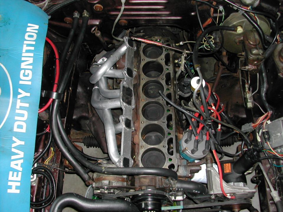 Inline-six engine with head removed showing six cylinder bores, silver header, and blue distributor cap in engine bay.