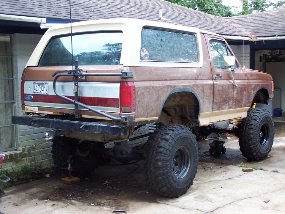 Rear three-quarter of lifted brown Ford Bronco with tan top and tube bumper on a driveway