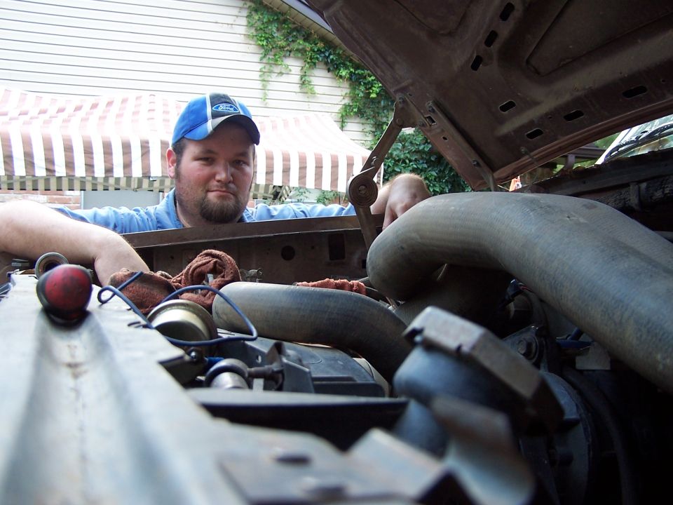 Man in blue Ford cap leaning over open engine bay showing radiator hoses and hood prop rod