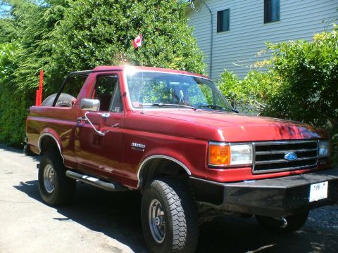 1989 Ford Bronco - 3/4-front view of a red Ford Bronco with the rear hardtop removed, parked on a driveway beside a white house and tall hedge. It sits on all…