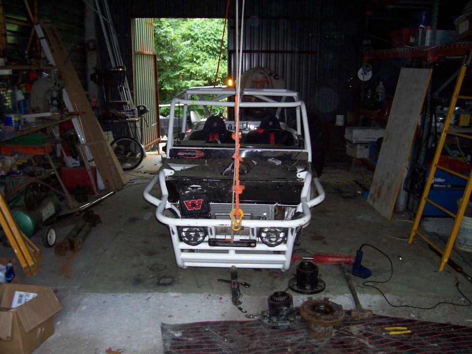 Custom off-road vehicle with white roll cage and bumper, being lifted by a hoist in a garage