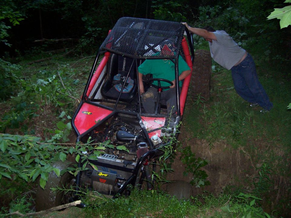 Red tube-chassis rock buggy nose-down in a muddy wooded ditch with two people working to free it