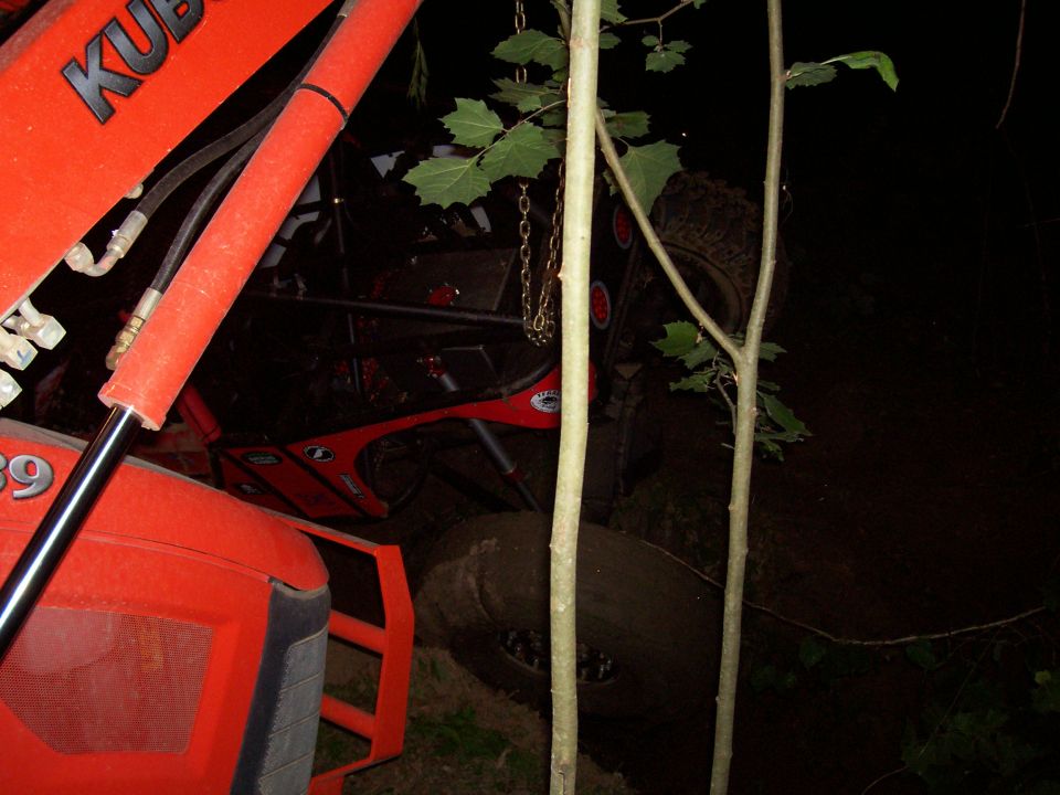 Night recovery, orange Kubota loader with chain over red off-road rig tipped on its side in brush