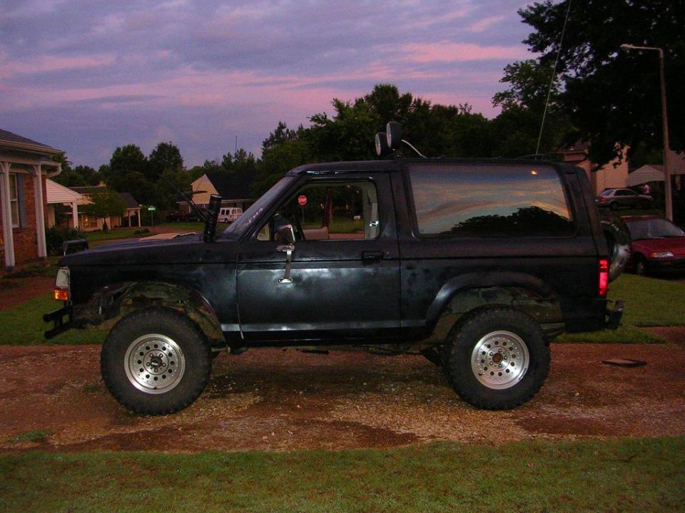 Side profile of lifted flat-black two-door Ford SUV on gravel drive at dusk with roof lights and mud tires
