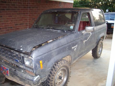 1986 Ford Bronco II - A dusty Ford Bronco II is parked in front of a brick wall, with another vehicle visible behind it.