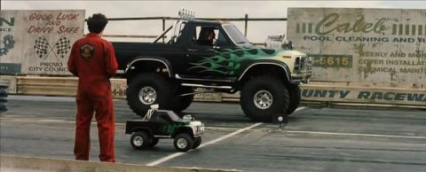1991 Ford Bronco - A large black Ford Bronco with green flame decals sits on a racetrack next to a much smaller replica, with a person in red overalls standin…