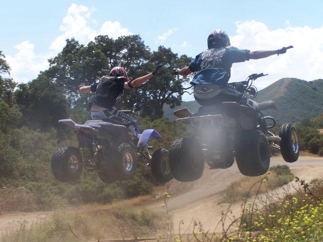 Two sport quads jumping mid-air on dirt trail, riders with arms out