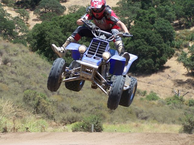 Rider on blue ATV airborne over a dirt track with trees and hills in background