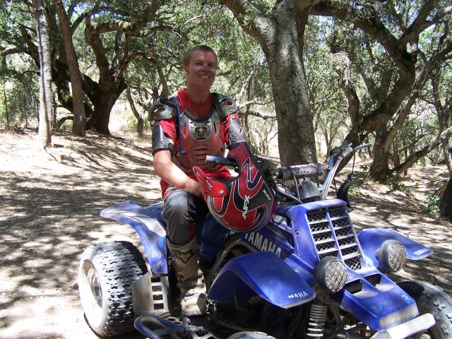 Man in chest protector seated on a blue Yamaha quad ATV on a wooded trail