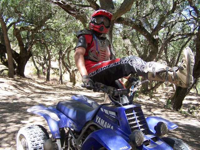 Rider in motocross gear sits on the handlebars of a blue Yamaha sport ATV on a wooded dirt trail