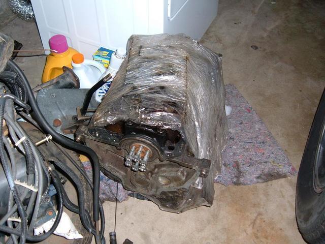 Engine block wrapped in plastic, with hoses and a tire visible