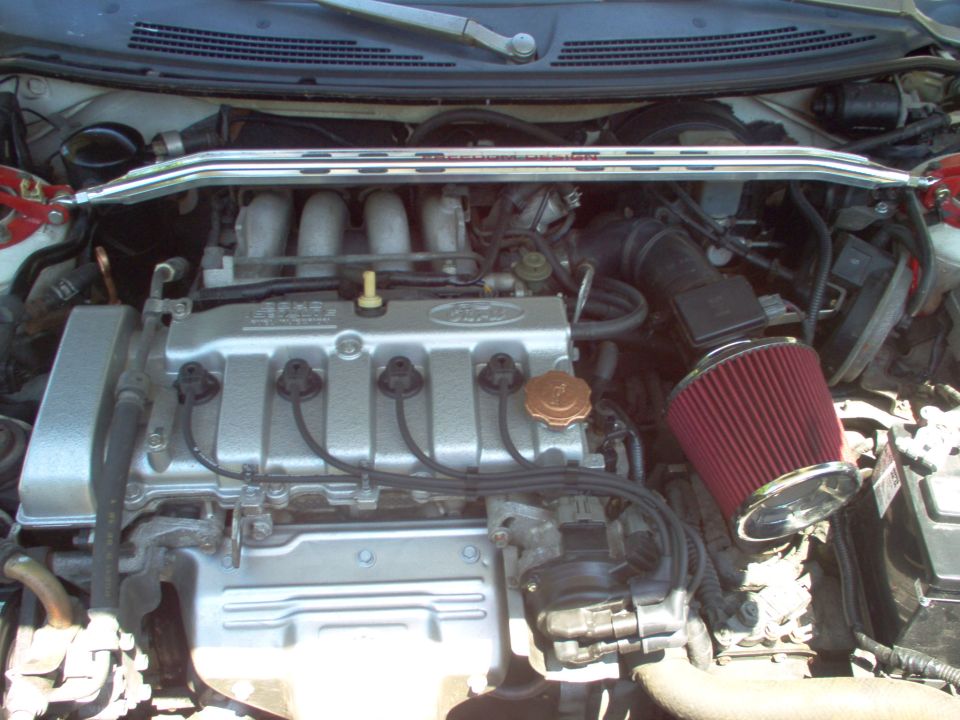 Engine bay of a car, showing the Ford engine, intake manifold, and a red aftermarket air filter. A strut bar is visible across the top