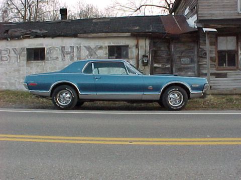 1968 Mercury Cougar - A blue 1968 Mercury Cougar is seen from the side, parked in front of a weathered building.