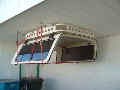 1990 Ford Bronco - A removed white fiberglass hardtop hoisted and stored under a carport roof, suspended by red ratchet straps and cable hoists anchored to th…