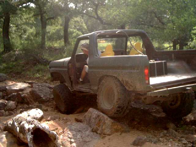 Mud-covered 1978 Ford Bronco off-roading over rocks, driver visible inside