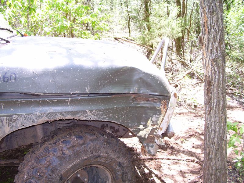 Damaged olive green vehicle fender and large mud-caked tire in wooded area. "360" is visible on the fender