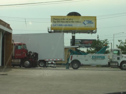 1992 Ford Bronco - A tow truck is positioned behind a semi-trailer truck, with a billboard visible in the background.