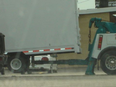 1992 Ford Bronco - A tow truck is hauling a trailer, with a white car partially visible underneath.