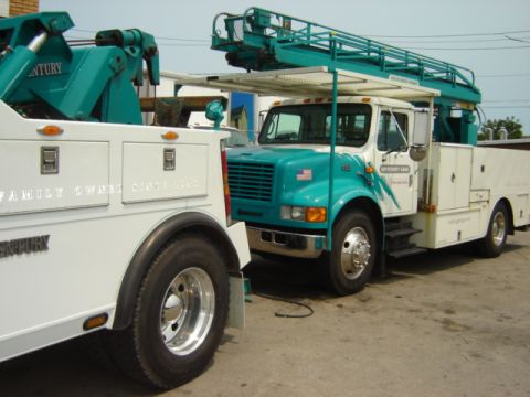 1992 Ford Bronco - A utility truck with a boom lift is parked next to a tow truck, with a water stain on the pavement.