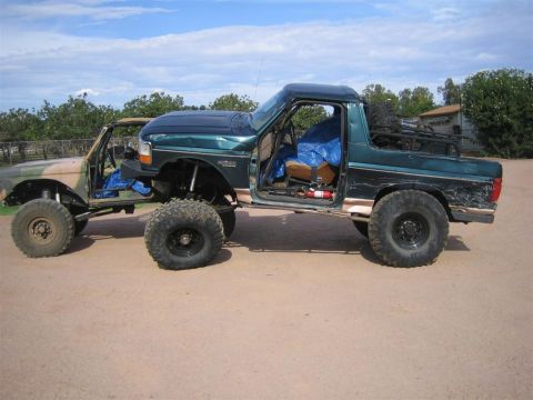 1994 Ford Bronco - Two custom Ford Broncos, one camouflaged, are parked outdoors on a dirt lot under a cloudy sky.