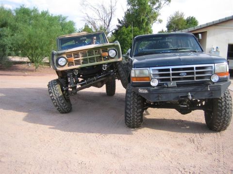 1994 Ford Bronco - Two lifted Broncos, one in camouflage and a dark blue 1994 model, are parked side-by-side on a dirt lot.