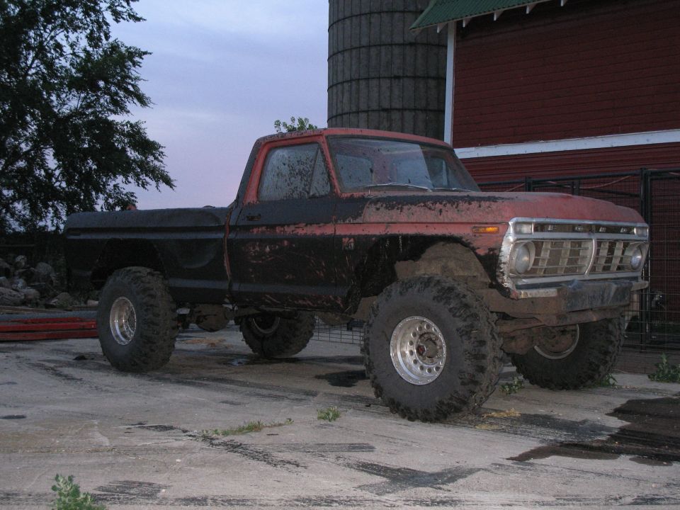 Lifted red and black Ford pickup covered in mud on big tires, parked by a red barn and silo at dusk