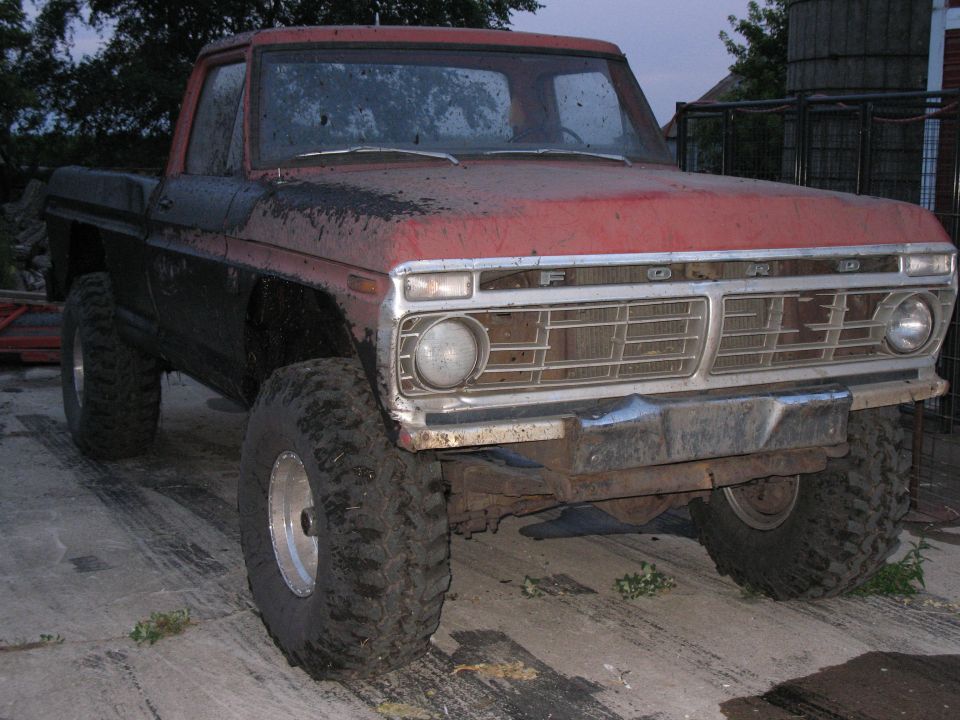 Lifted red and black Ford pickup on big mud tires, chrome grille, parked on concrete at dusk